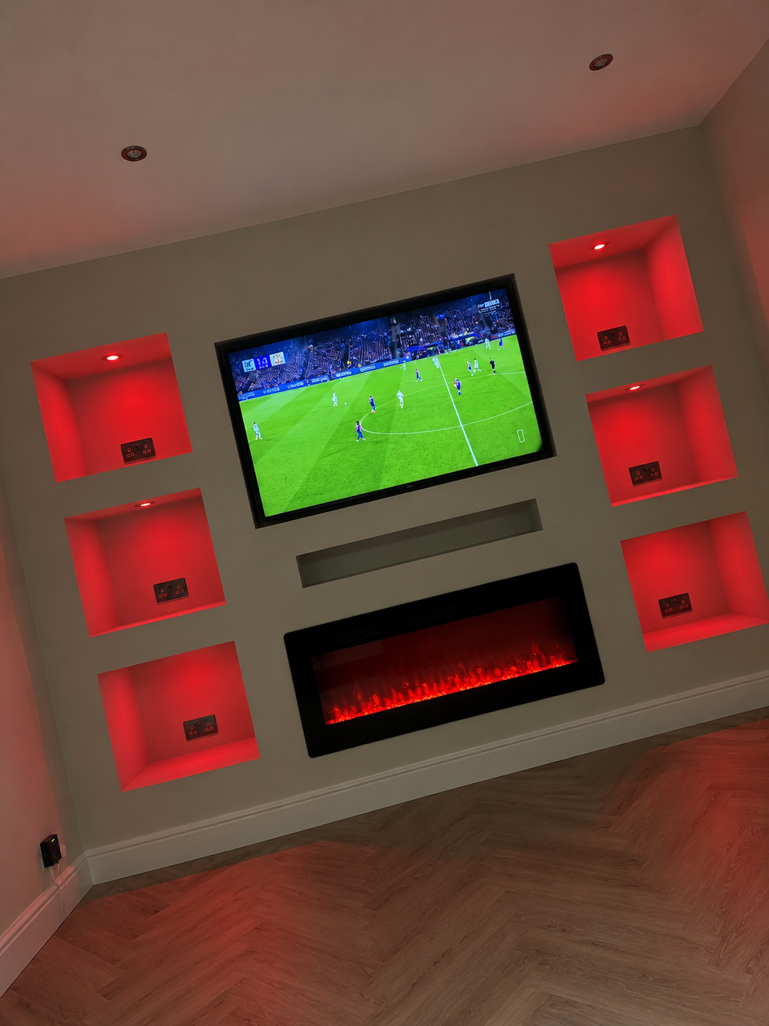 A modern built-in media wall featuring a recessed flat-screen TV playing a soccer game, a linear electric fireplace, and six square display niches illuminated with red LED lighting.